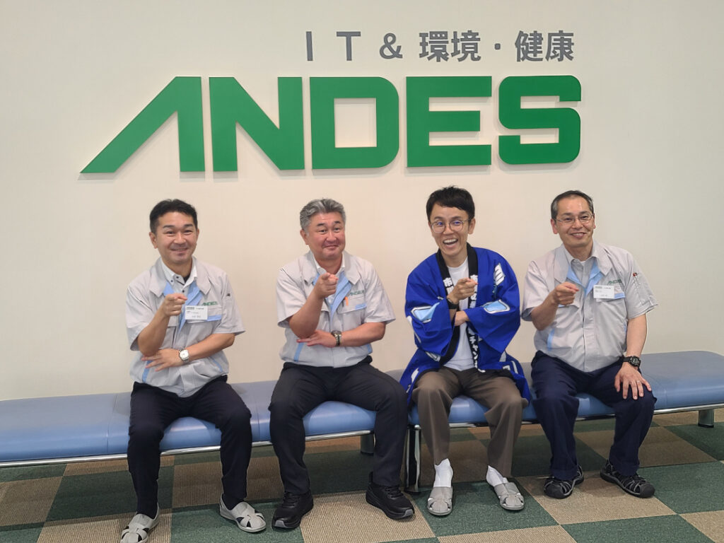アンデス電気本社エントランスで、当社の役職員3名と記念撮影する『女と男』の市川さん(右から2番目、青い法被姿)